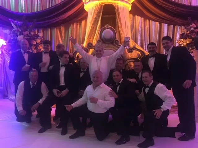 A group posing beside a Mardi Gras throne under purple and gold drapes
